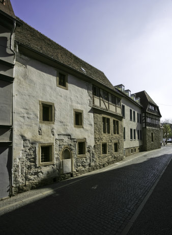 Foto: Steinernes Haus Außenansicht © TLDA, W. Streitberger Eine mit Kopfstein gepflasterte Straße im Sonnenlicht. Eine Reihe unterschiedlich alter Häuser ist zu sehen, eines mit Fachwerk, eines aus grob verputztem Mauerwerk, ein zeitgenössisches Gebäude.